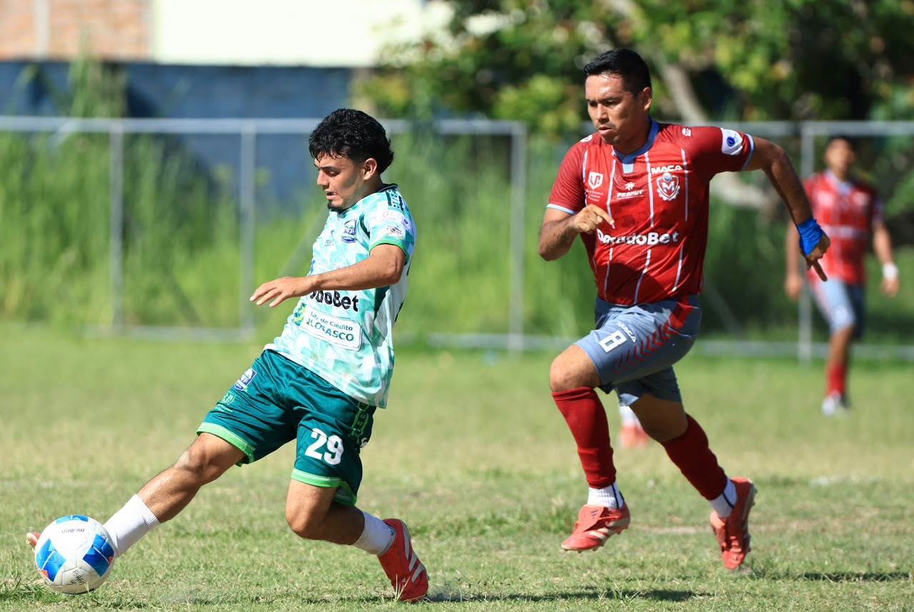 Jugador de verde controla el balón perseguido por un rival durante un partido